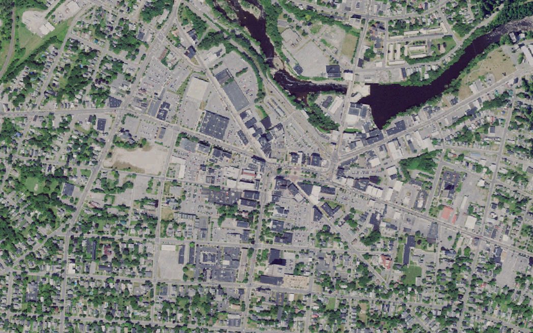 Aerial photograph of downtown Watertown, New York, showing the Black River,
              Public Square, dense blocks of buildings, tree cover and parking lots.